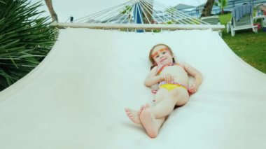 Mayolu ve iki örgülü saçı olan küçük bir kız plaj hamağı ve otel odasında güneşleniyor..