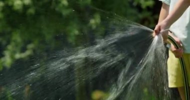 Bir kızın elleri bahçe hortumunu tutarken, sıcak bir günde su sıkarken. Su damlacıkları güneş ışığında parıldar, yaz sıcağında serinlemenin neşesini ve ferahlığını yakalar. Sahne