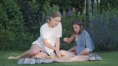 İki arkadaş arka bahçede bir battaniyeye oturmuş piknik sırasında mutlu bir şekilde pizza yerken görülüyor. Sahne, açık hava yemeğinin rahat ve eğlenceli atmosferini iyi bir arkadaşla yakalıyor..