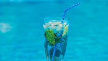 Mavi pipetli, ferahlatıcı bir içecek, nane yaprakları ve buzla dolu yakın plan bir bardak. Cam canlı bir mavi su arka planına dayanır, serinlik ve ferahlık hissi uyandırır.