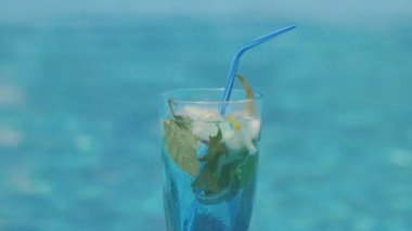 Mavi pipetli, ferahlatıcı bir içecek, nane yaprakları ve buzla dolu yakın plan bir bardak. Cam canlı bir mavi su arka planına dayanır, serinlik ve ferahlık hissi uyandırır.