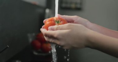 Kadınların elleri, mutfak musluğundan gelen su püskürtülerinin altında olgun kırmızı bir domates yıkar..