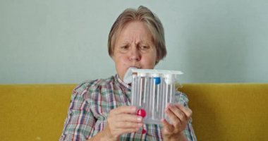 Yaşlı bir kadın, akciğer kapasitesini ölçmek ve solunum kaslarını güçlendirmek için tasarlanmış bir cihaz olan spirometreye üfleyerek sağlık egzersizi yapıyor. Bu aktivite onun rutininin bir parçası.
