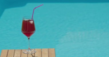 Soğuk bir kokteyl ile dolu bir bardak, parlak bir yaz gününde bir havuzun su yüzeyine konur. Güneş ışığı sudan yansıyarak, içeceğin canlı renklerini yükseltir ve bir...