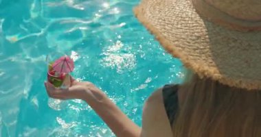 Şık bir şapka takan, elinde canlandırıcı bir içecek tutan genç bir kadın havuzun kenarında oturuyor. Parlak güneş ışığı, sıcak bir içecekten hoşlanırken onun rahat ve memnun yüz ifadesini aydınlatır.