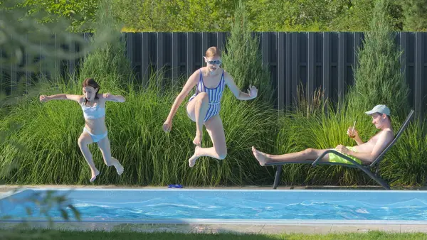 stock image Two teenage girls, one in a striped swimsuit and sunglasses and the other in a light blue swimsuit, are captured mid-air as they jump into a backyard pool. A man is relaxing on a lounge chair beside