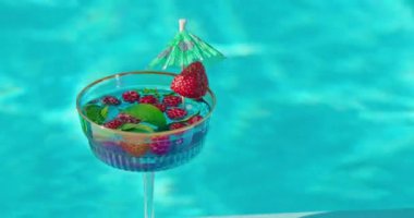 Taze ahududu ve çilekle süslenmiş ferahlatıcı bir içecekle dolu bir kokteyl bardağı havuz suyu yüzeyinin zeminine yerleştirilir. Canlı kırmızı böğürtlenler ve açık mavi