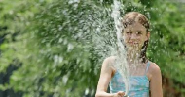 Mutlu bir kız bahçe hortumuyla oynarken yakalanıyor, neşeyle su püskürtüyor ve gülüyor. Sahne, suyun ferahlatıcı serinliğini ve çocukluğun sıcak ve kaygısız anlarını vurgular.