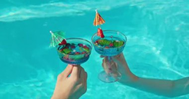 Serinletici içeceklerle dolu zarif bardakları tutan iki el havuz suyu yüzeyine kaldırılır. Parlak, renkli içecekler ve berrak mavi su canlı ve şık bir yaz manzarası yaratır.