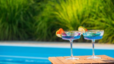Mavi kokteyllerle dolu iki sofistike bardak, her biri çilek ve limonla süslenmiş, ahşap bir masada dinleniyor. Canlı yeşil yapraklar ve arka plandaki açık mavi havuz.