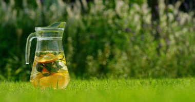 Limon dilimleri ve nane şekerleriyle doldurulmuş ferahlatıcı limonata dolu cam sürahi canlı yeşil bir çimenlikte duruyor. Arkaplan özellikleri bulanık yeşillik, davet ve artırma