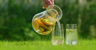Dondurulmuş limonatayı taze limon dilimleri ve cam sürahide bulunan nane yapraklarıyla doldurup iki uzun bardağa dolduran bir elin yakın çekimi. Sahne canlı yeşil bir çimenlikte serinliyor.