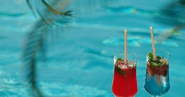 İki uzun kokteyl bardağı, biri kırmızı içecek, diğeri mavi içecek, havuzun yanında otur. Her içecek taze nane yaprakları ve meyvelerle süslenir. Palmiye yapraklarının yansıması