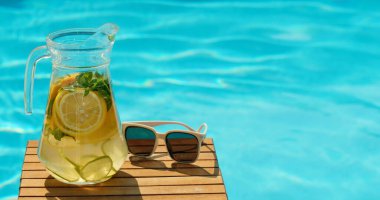 Sürahi dolusu soğuk su, limon dilimleri ve taze nane yaprakları şık beyaz güneş gözlüklerinin yanındaki ahşap masada duruyor. Arka planda açık, davetkar bir mavi havuz var.