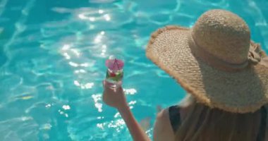 Şık bir şapka takan, elinde canlandırıcı bir içecek tutan genç bir kadın havuzun kenarında oturuyor. Parlak güneş ışığı sıcak bir parıltı saçar, rahat duruşunu vurgular ve serin bir içkinin tadını çıkarır.