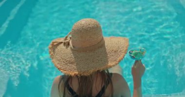 Havuzda dinlenirken elinde bardak tutan genç bir kadının arka görüntüsü. Parlak güneş ışığı sudan yansıyarak ılık bir yaz gününde içkisinin tadını çıkarırken huzurlu ve ferahlatıcı bir atmosfer yaratır.