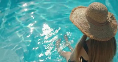 Şapka takan genç bir kadın havuzun kenarında oturuyor, ayakları suda sallanıyor. Güneş ışığı, suyun ferahlatıcı serinliğinin tadını çıkarırken onun rahat ve memnun ifadesini vurgular.