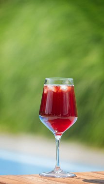 Kırmızı buzlu bir içecek içeren uzun bir bardak, yemyeşil yeşillik bir zemin üzerine kurulmuş, ahşap bir yüzeye oturur. İçkinin canlı kırmızısı yeşillikle güzel bir zıtlık yaratıyor.