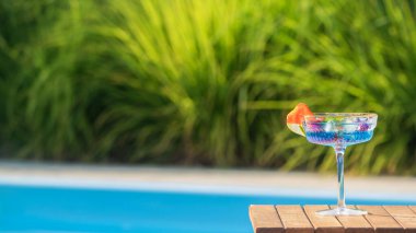 Çilek dilimi ve limon dilimiyle süslenmiş bir bardak mavi kokteyl ahşap bir masada duruyor. Arka plandaki berrak havuz ve yemyeşil yeşillik canlandırıcı ve canlı hissi arttırır.