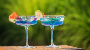 Her biri bir dilim çilek ve bir limon dilimiyle süslenmiş iki bardak mavi kokteyl ahşap bir masaya yerleştirilir. Canlı yeşil arka plan canlandırıcı ve davetkar doğasını arttırır.