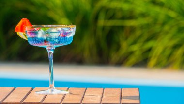 Bir bardak mavi kokteyl ahşap bir yüzeye yerleştirilir, çilek dilimi ve limon dilimi ile süslenir. Canlı yeşil arka plan içkinin ferahlatıcı ve davetkar görünümünü vurguluyor.