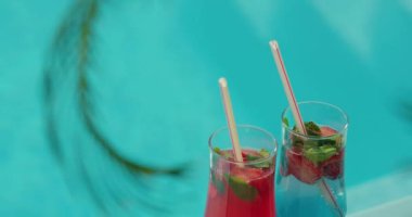 İki ferahlatıcı soğuk kokteyl bir havuzun kenarında oturur ve palmiye yaprakları ön planda yavaşça sallanır. İçkilerin canlı renkleri ve tropikal ortam davetkar bir sahne yaratıyor.