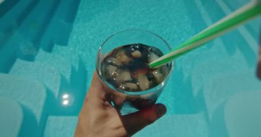 Birinci şahıs bakış açısına göre, bir adam havuza bakarken ferahlatıcı bir içecekle buz karıştırırken görülüyor. Arka plandaki soğuk içecek ve köpüklü su rahatlatıcı ve canlandırıcı bir şey yaratıyor.