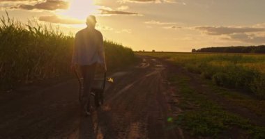 Botlu bir çiftçi mısır tarlasının yanındaki toprak yolda yürüyor, arka planda güneş batarken el arabasını itiyor. Görüntü çiftçinin siluetini yakalıyor. Sıcak parıltıya karşı.
