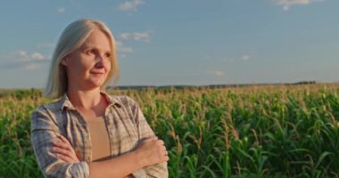 Huzurlu orta yaşlı bir kadın, sarı saçları altın güneş ışığıyla hafifçe aydınlatılmış, canlı bir mısır tarlasının önünde duruyor. Sıradan bir ekose gömlek giyiyor, kollarını kavuşturup jest yapıyor.