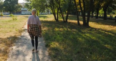 Orta yaşlı bir kadın, görünüşte yorgun ama memnun, sabahın erken saatlerinde bir stadyumun yakınında yürüyor. Rahat duruşu ve yüzündeki sakin ifade tatmin edici ifadeyi yansıtıyor.