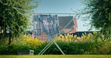 A scene of freshly washed clothes drying on a foldable laundry rack in a sunny backyard. The laundry rack is placed on a green lawn, surrounded by lush greenery and trees. A storage basket sits on the clipart