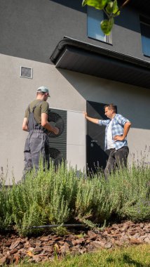 A technician in overalls discusses the setup and maintenance of a heat pump with a homeowner, standing next to the outdoor unit on the side of the house. The conversation takes place in a well clipart