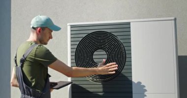 İş yerinde çalışan bir teknisyen, modern bir evin yanına kurulmuş açık hava ısı pompası ünitesini ayarlarken görülüyor. Bir elinde tablet tutarken diğer elinde pompaların işlevselliğini kontrol ediyor.