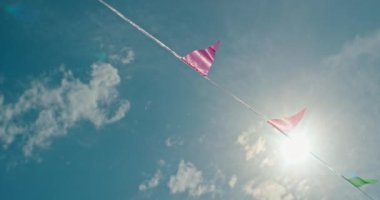 Kağıt bayraklardan yapılmış renkli bir çelenk rüzgarda hafifçe sallanıyor, parlak mavi gökyüzünün arka planında güneş parlıyor. Bayrakların hareketi ve canlı bir gökyüzü yaratır.