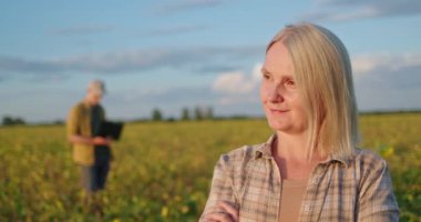 Kararlı bir kadın lider bir tarlada dikilirken görülüyor, kollarını kavuştururken kendine güven yayıyor. Arkasında, bir adam bilgisayarına odaklanmış modern teknolojinin entegrasyonunu sembolize ediyor.