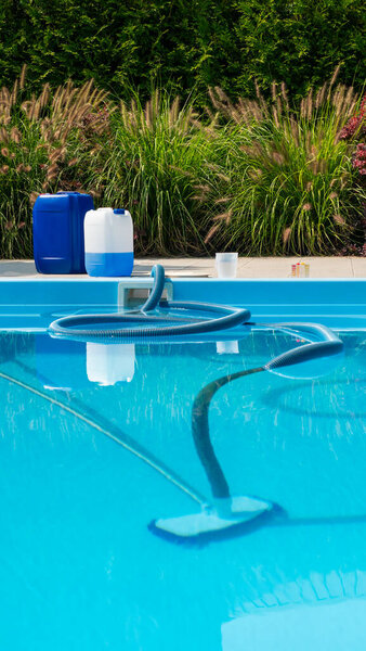 Шланг для очистки бассейна погружается в чистую голубую воду бассейна, с большими химическими контейнерами и набором для тестирования воды, видимым на поверхности бассейна. Сцена украшена яркой зеленью