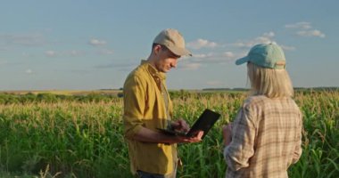 Bir adam ve bir kadın, her ikisi de çiftçi, geniş bir mısır tarlasının önünde dikilip sohbet ediyorlar. Adam bir dizüstü bilgisayara odaklanmış, onların tarım uygulamalarını desteklemek için modern teknolojiyi kullanıyor. Ayyaşlar.