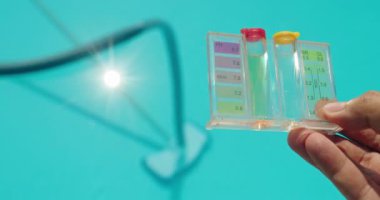 Bir el, parlak mavi bir yüzme havuzunun zeminine karşı pH ve klor seviyelerini gösteren bir havuz suyu test kiti tutuyor. Güneş ışığı havuz suyundan yansıyarak ışıl ışıl bir etki yaratıyor.