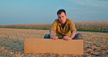 Bir adam elinde boş bir karton levhayla bir tarlada oturuyor. O, sarı bir gömlek giymektedir ve kırsal manzara onun arkasında açık mavi bir gökyüzünün altında uzanır. Bu resim mükemmel.