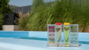 PH ve klor seviyelerini gösteren renkli sıvılarla dolu şeffaf tüpler içeren havuz suyu test setinin detaylı bir görüntüsü. Havuz kenarında konumlanmış, net değerlere odaklanmış durumda.