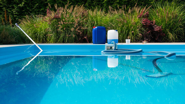 На этом изображении изображен прозрачный голубой бассейн с моющим шлангом, частично погруженным в воду. На берегу бассейна находятся большие контейнеры для химикатов и комплект для анализа воды.