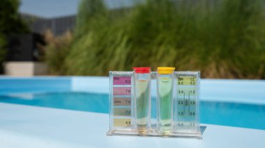 Bir havuz suyu test kitinin yakın çekiminde pH ve klor seviyeleri, renkli göstergelerle dolu şeffaf tüplerle gösterilir. Ekipman havuzun kenarına yerleştirildi, arka plan yumuşak.