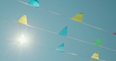 Rüzgarda dalgalanıp duran kağıt bayraklar güneş ışığıyla aydınlatılarak açık hava kutlaması için şenlikli bir ortam yaratıyor. Parlak mavi gökyüzü canlı bir zemin görevi görüyor, neşeyi arttırıyor.