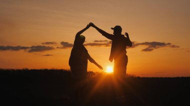 İki çiftçi bir tarlada neşeyle dans ediyor. Siluetler güneşin battığı gökyüzüne karşı.
