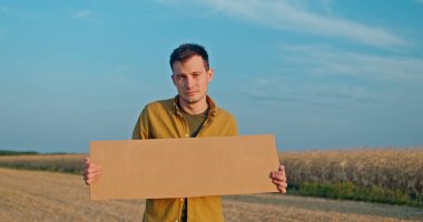 Bir adam geniş bir alanda elinde boş bir karton levhayla duruyor. Sarı bir gömlek giymişti. Geniş mavi gökyüzü ile çelişiyordu. Görüntüyü çeşitli temalar için ideal yapıyordu.