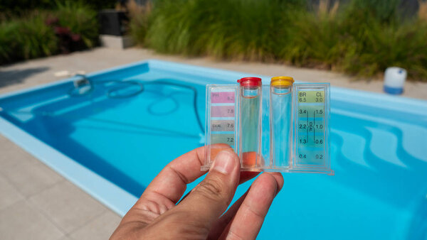 Крупный план руки, держащей комплект для тестирования воды над прозрачным голубым бассейном, с оборудованием для очистки бассейна на заднем плане. Сцена подчеркивает важность сохранения воды
