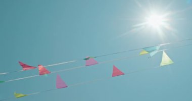 Renkli flama bayrakları hafifçe dalgalanır berrak, parlak bir gökyüzünün arka planına güneş ışıl ışıl parlarken. Canlı bayraklar, yeşil, mavi ve sarı tonlarında, sahneye neşeli bir dokunuş ekler.