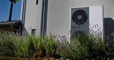 Çağdaş bir evin duvarına, lavanta bitkileriyle çevrili bir açık hava ısı pompası monte edilir. Evin ince çizgileri arka planda görülebilirken, yakındaki fabrika da görülebiliyor.