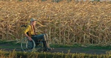 Tekerlekli sandalyedeki genç bir çiftçi güneşli bir günde açık mavi bir gökyüzünün altında mısır tarlası boyunca hareket eder. Sahne, esneklik, sakatlık ve tarım hayatıyla ilgili konuları vurguluyor.