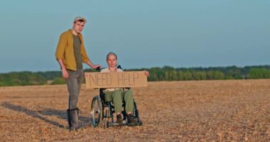 Genç bir adam, tekerlekli sandalyedeki yaşlı bir kadının yanında duruyor. Elinde kuru bir tarlada 